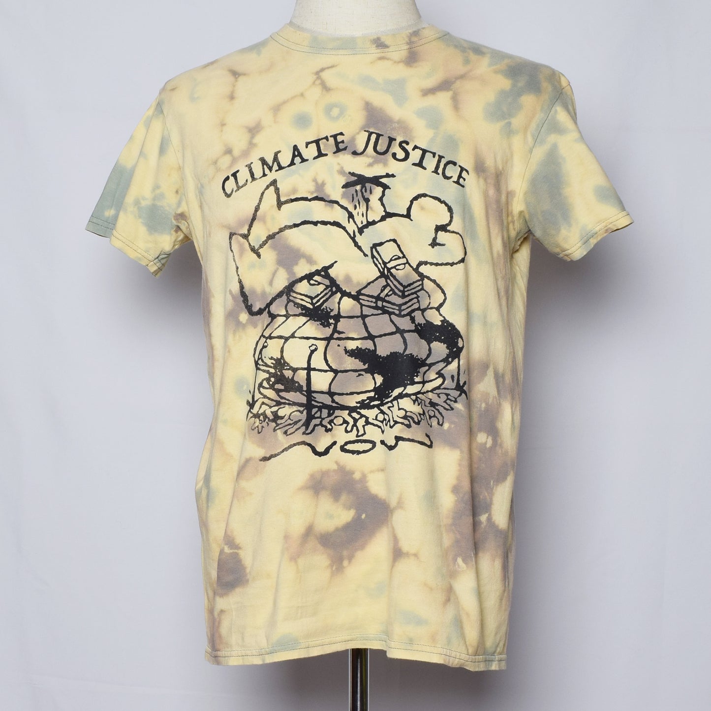 イエローとグリーンのタイダイTシャツにClimate Justice NOWの文字と、多くの人と地球が戦闘機と札束を持った人間に押しつぶされているグラフィックがプリントされている