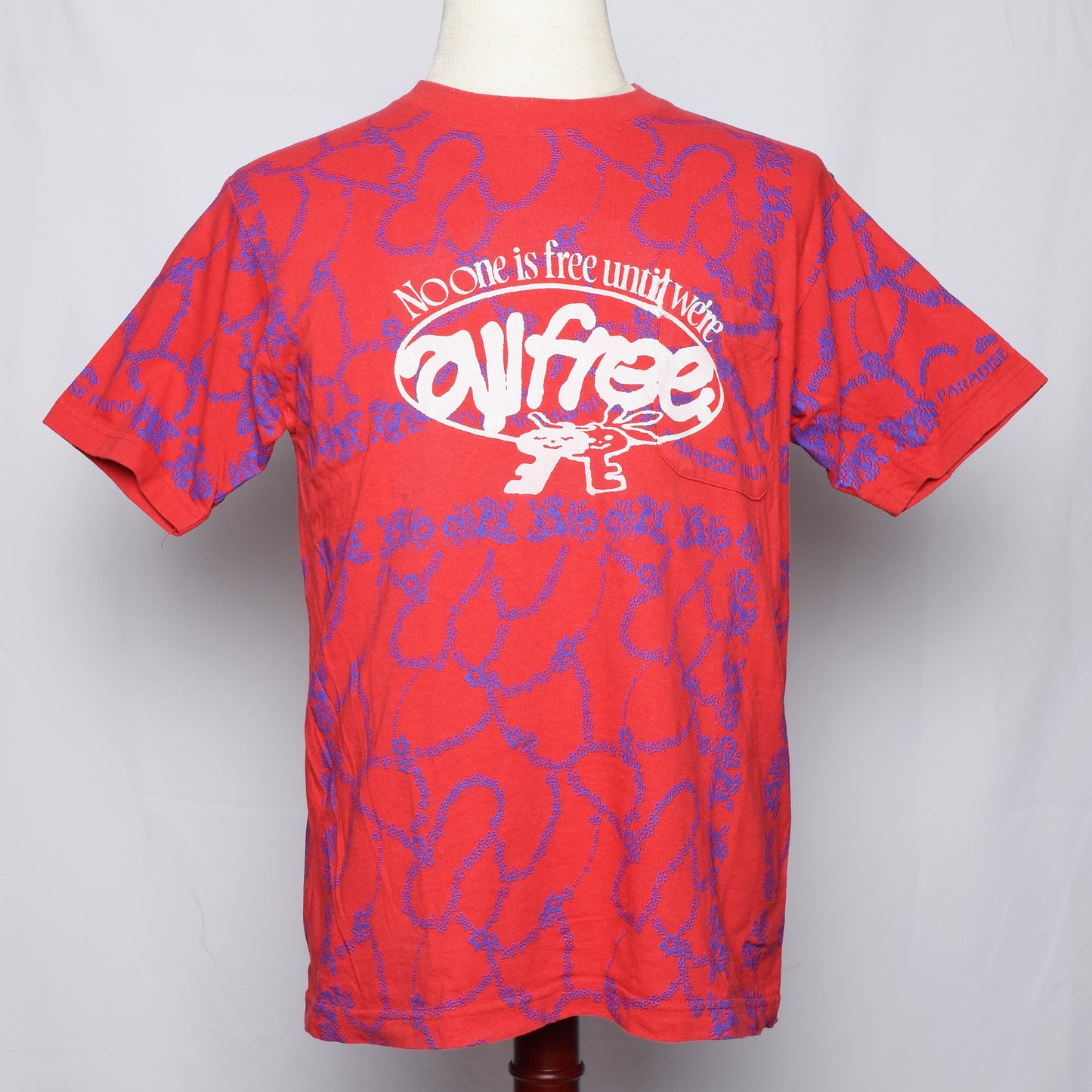 青い細かいライン状の花の柄の赤いTシャツにNo one is free until we are all freeの文字と鍵のキャラクターがプリントされている