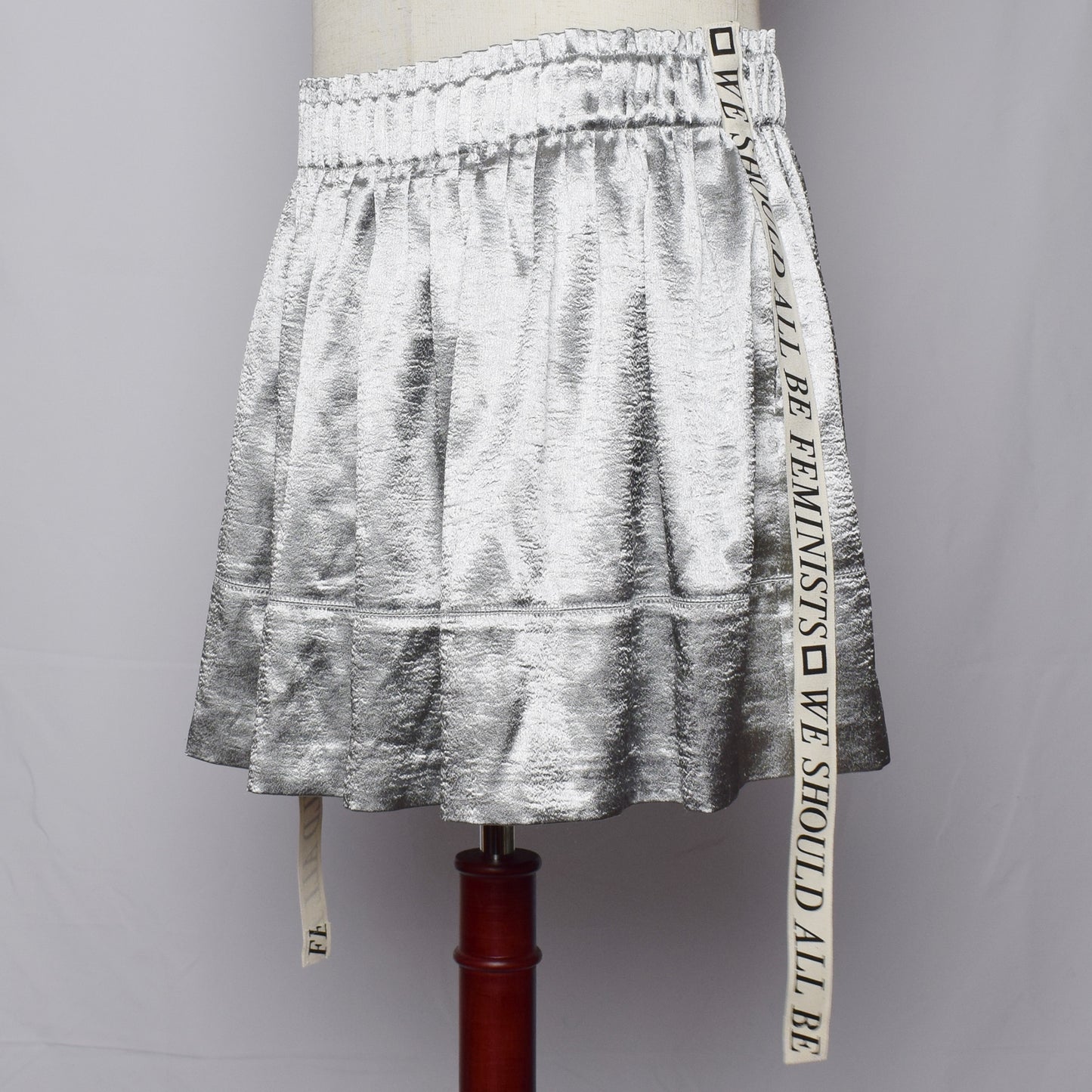 ウェストに細かいプリーツの入ったシルバーのショートパンツのサイドに、We should all be Feministsとプリントされたリボンが縫い付けられている。