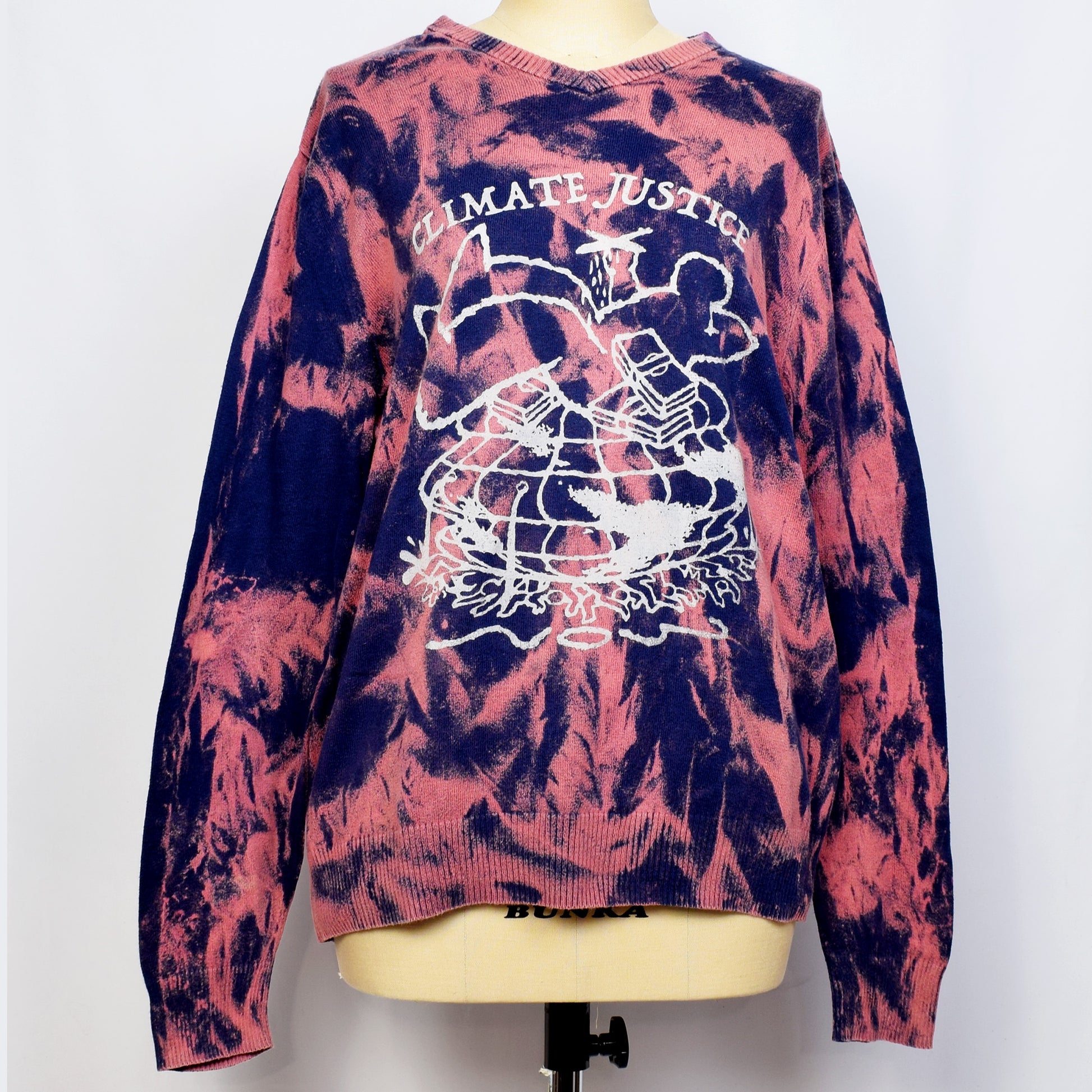 ネイビーとピンクのタイダイのセーターにClimate Justiceと戦闘機を持った一人の大きい人間と札束が大勢の人と地球を押し潰しているイラストがプリントされている。