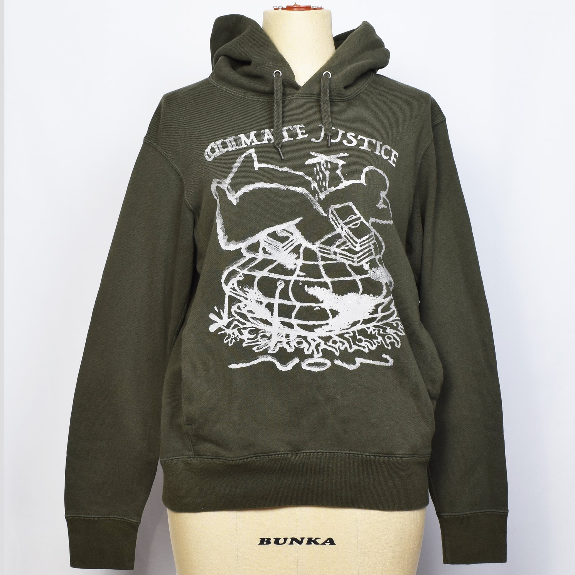 オリーブ色のプルオーバーパーカーにClimate Justiceと戦闘機を持った一人の大きい人間と札束が大勢の人と地球を押し潰しているイラストがプリントされている。