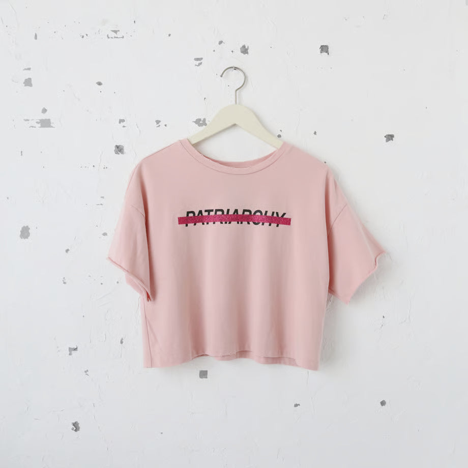 ピンク色の短めのTシャツにPatriarchyの文字とそれを打ち消すようにピンクのラメのキラキラでプリントされている。ダメージ加工あり。