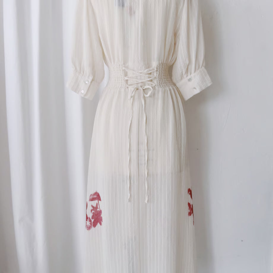 シアー素材のオフホワイトの七分袖のワンピース。謎の動物が手を繋いでいるシルエットのグラフィックが赤い色で数箇所プリントされている。
