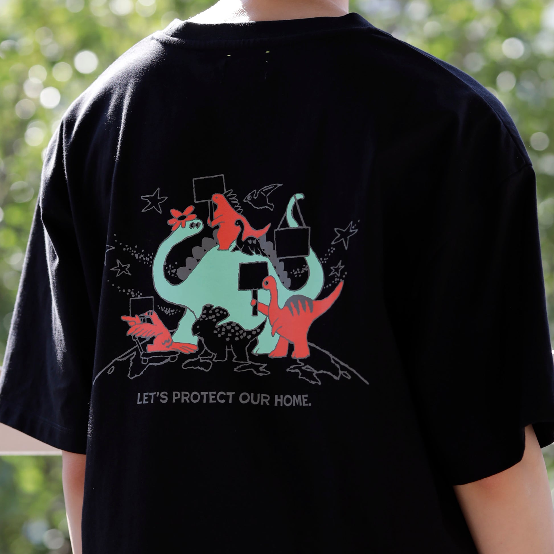 黒いTシャツのバックプリントとして、様々な種類の恐竜たちがプラカードを持って行進しているレッドとミントグリーンのイラストがあり、その下に、「LET'S PROTECT OUR HOME.」の文字がある。
