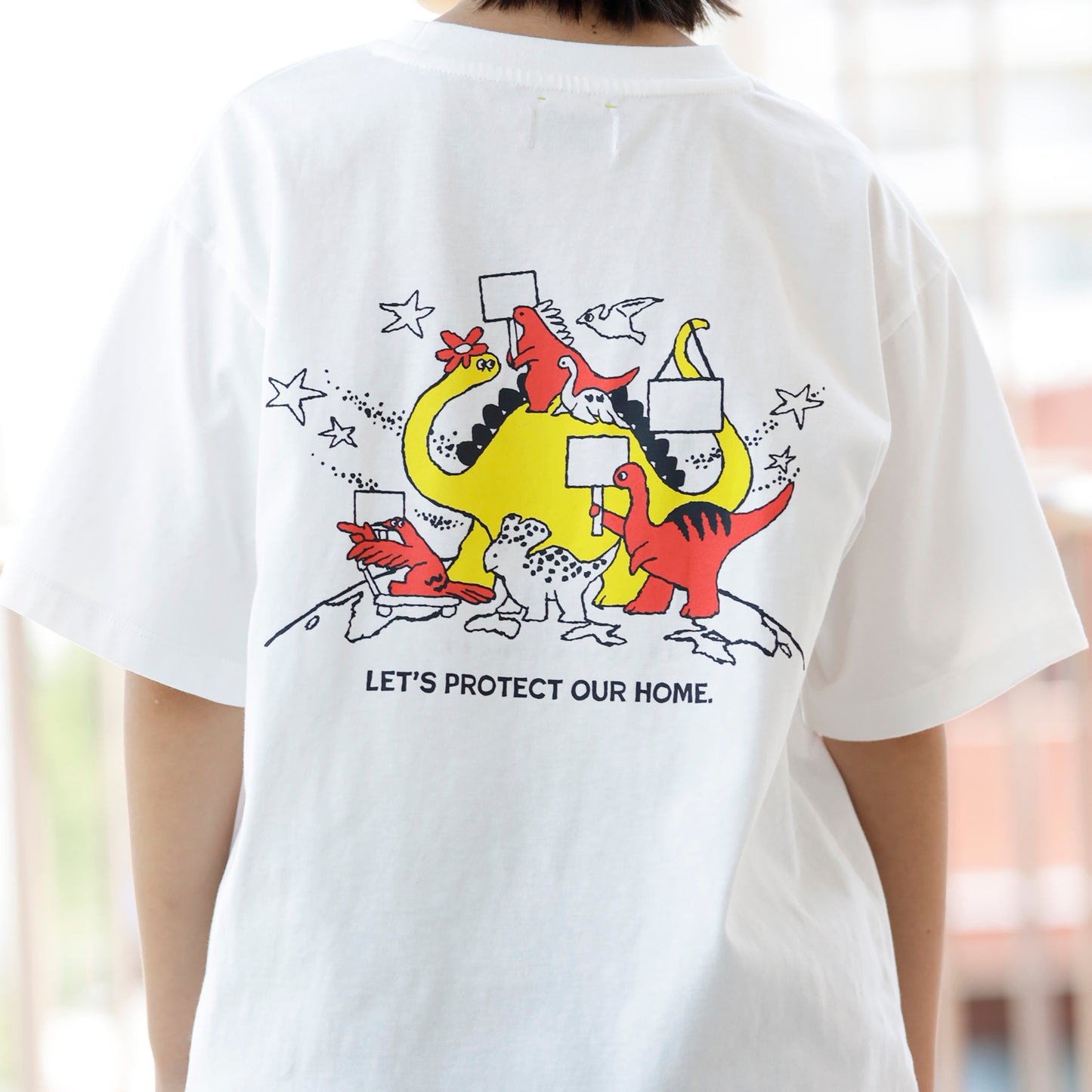 白いTシャツのバックプリントとして、様々な種類の恐竜たちがプラカードを持って行進しているレッドとイエローのイラストがあり、その下に、「LET'S PROTECT OUR HOME.」も文字がある。