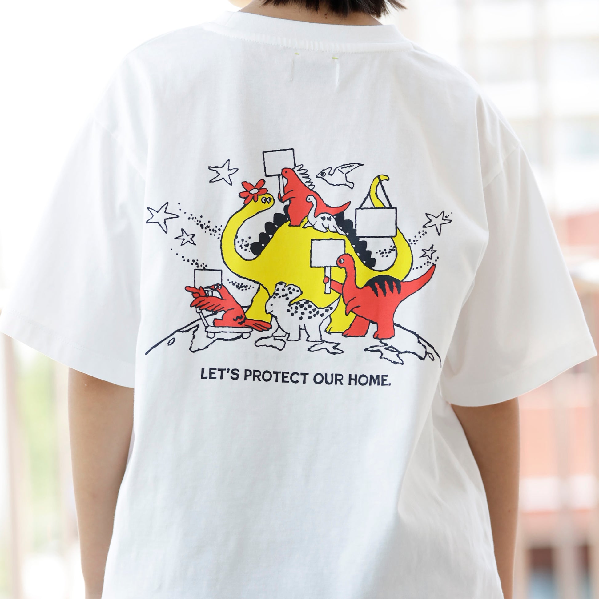 白いTシャツのバックプリントとして、様々な種類の恐竜たちがプラカードを持って行進しているレッドとイエローのイラストがあり、その下に、「LET'S PROTECT OUR HOME.」も文字がある。